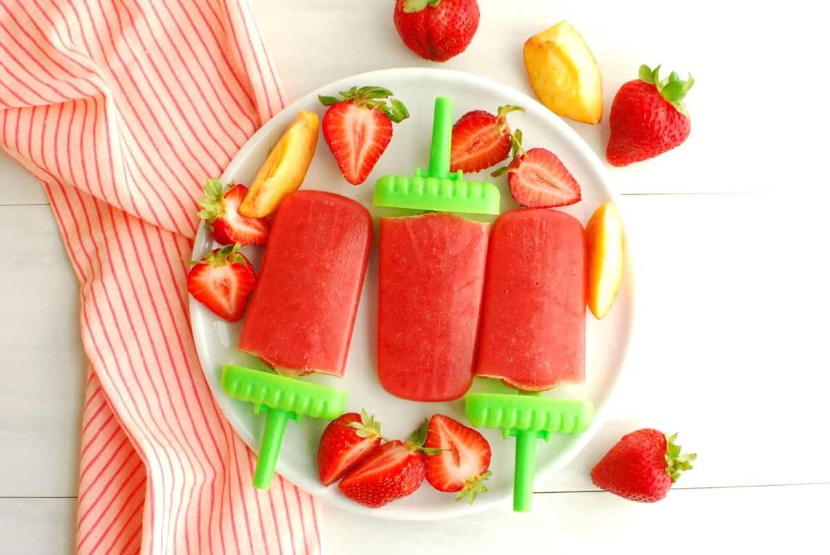 Three strawberry peach popsicles next to some fresh cut peaches and strawberries.