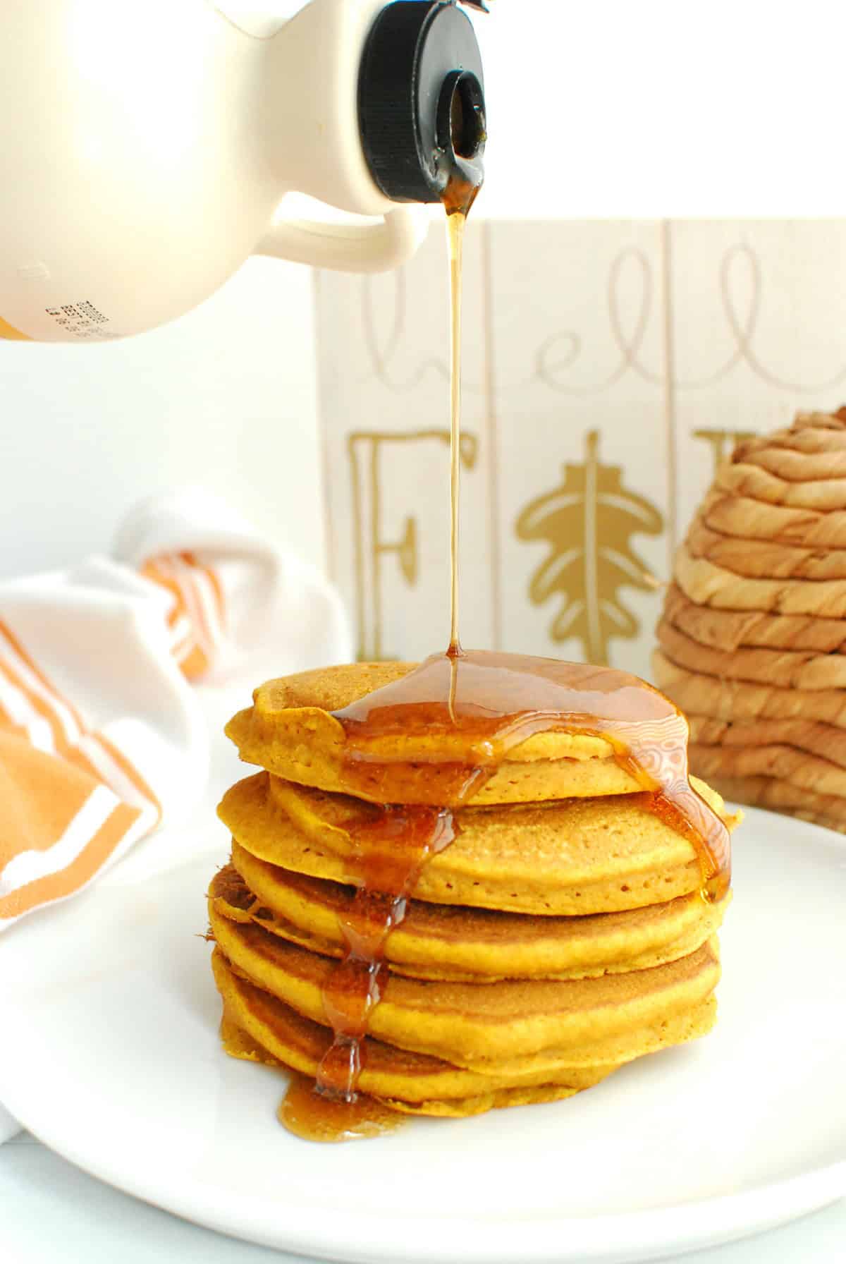 A plate full of whole wheat pumpkin pancakes with maple syrup being drizzled on top.