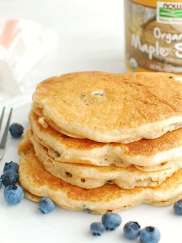 A stack of four blueberry Greek yogurt pancakes on a white plate, next to a fork and some scattered blueberries.