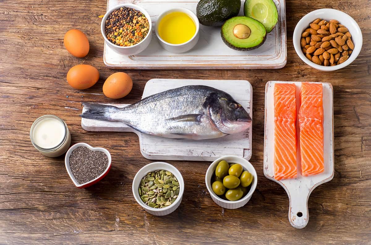 Assorted food sources of fat on a wooden table including fish, nuts, seeds, and avocado.