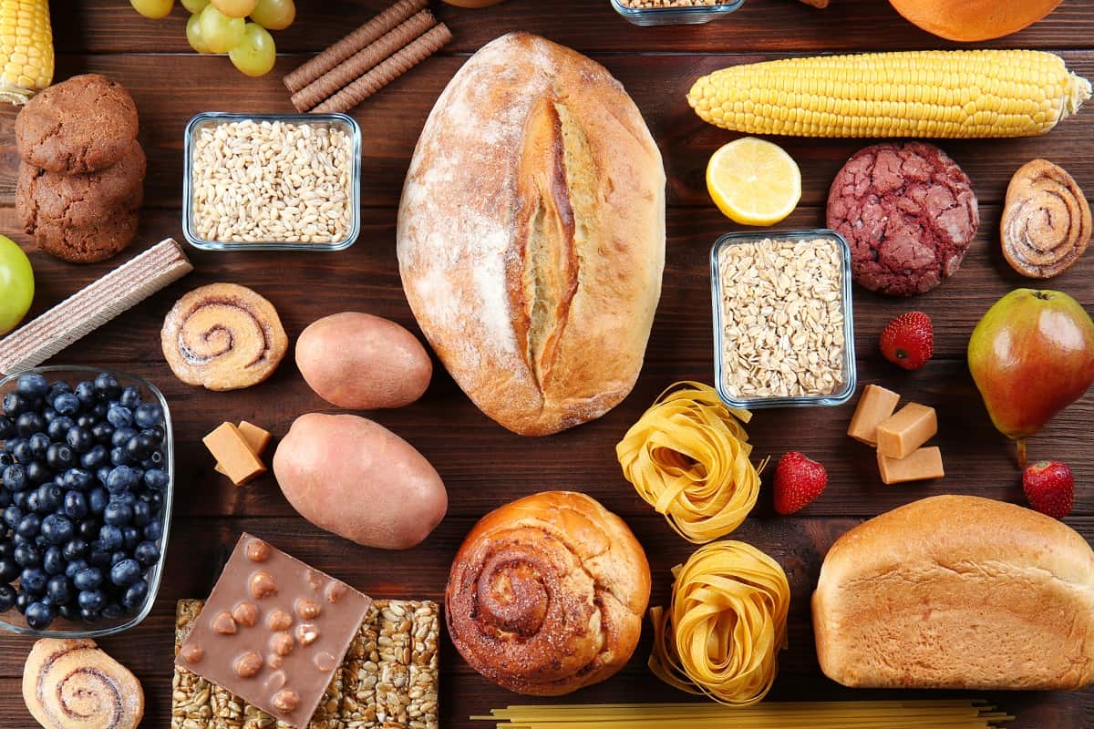Assorted carbohydrate rich foods on a wooden table like bread, blueberries, corn, and pasta.