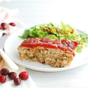 A slice of cranberry chicken meatloaf on a white plate next to a napkin and scattered cranberries.