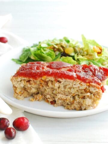 A slice of cranberry chicken meatloaf on a white plate next to a napkin and scattered cranberries.