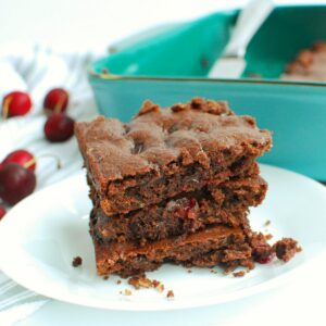 Three chocolate cherry brownies on a plate next to a pan of brownies and some scattered fresh cherries.