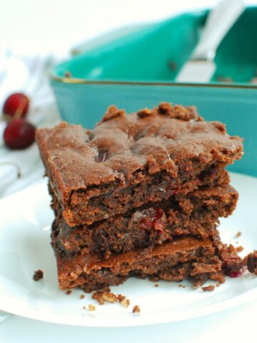 Three chocolate cherry brownies on a plate next to a pan of brownies and some scattered fresh cherries.