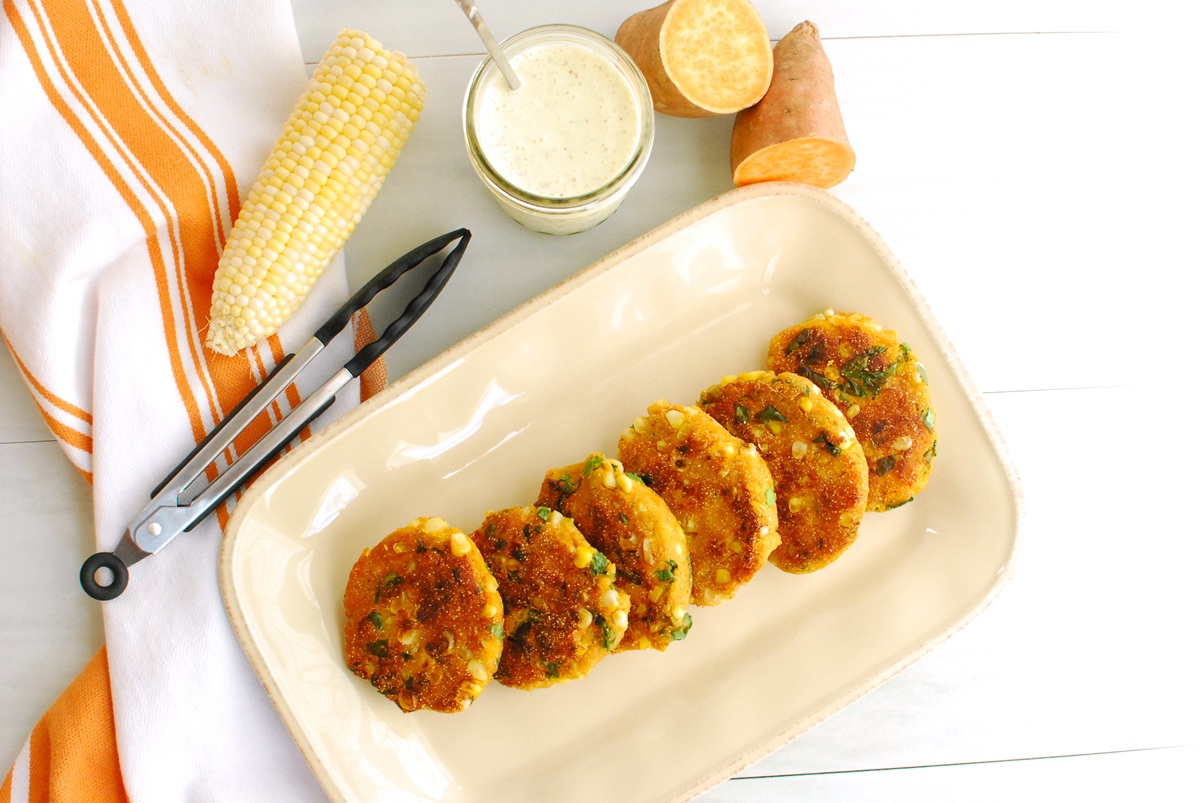Six corn sweet potato fritters on a plate next to some tons and sweet corn.