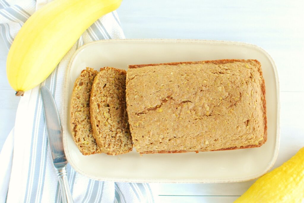 Overhead shot of a loaf of yellow squash bread next to two fresh picked yellow squash and a napkin.