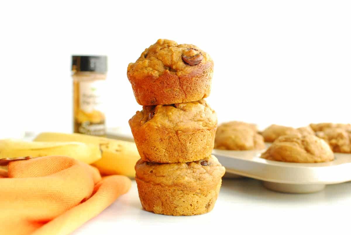 Three pumpkin banana muffins stacked on top of each other, with a muffin tin and pumpkin spice in the background.
