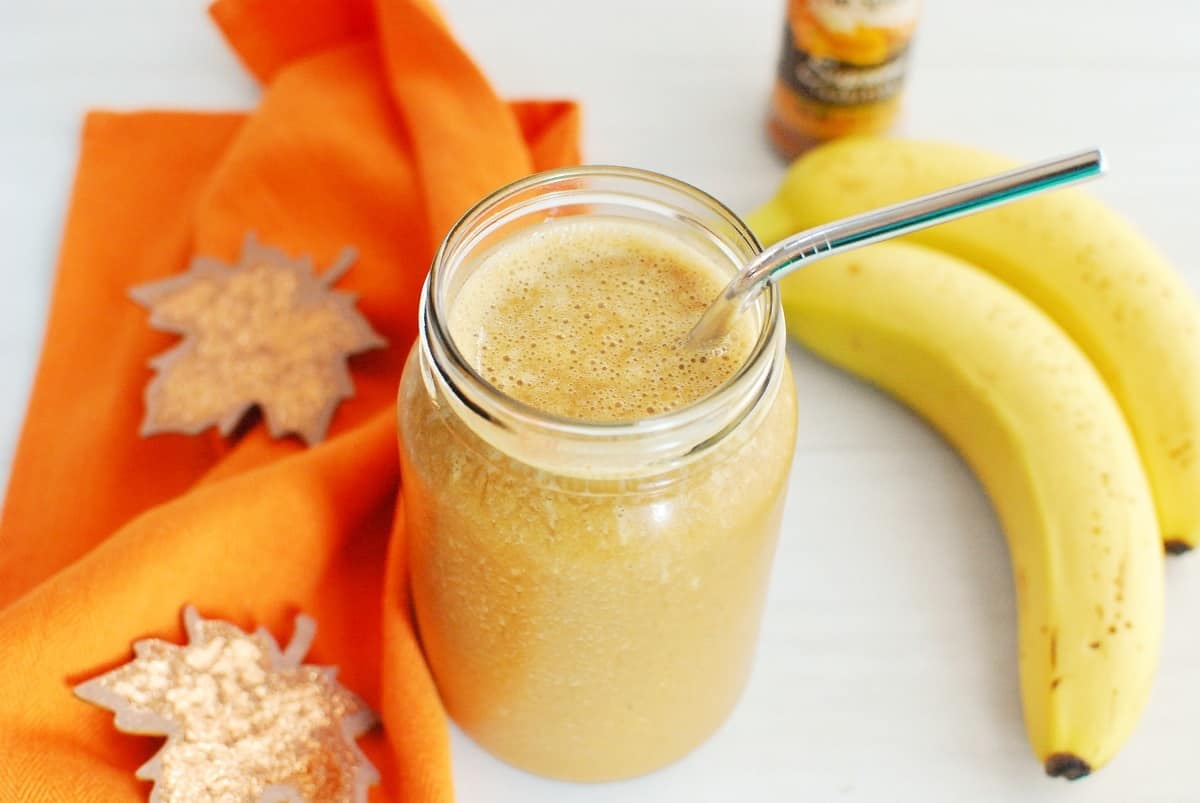 A pumpkin coffee smoothie with a straw next to bananas, pumpkin spice, and an orange napkin.