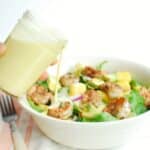 A woman holding a jar of creamy citrus salad dressing, pouring it onto a shrimp salad.