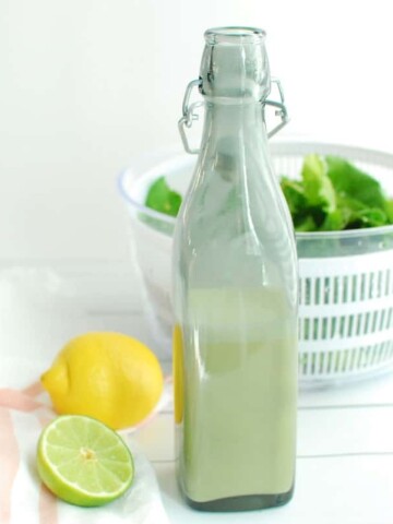 A salad dressing bottle filled with creamy citrus dressing, next to a lemon, lime, and leafy greens.