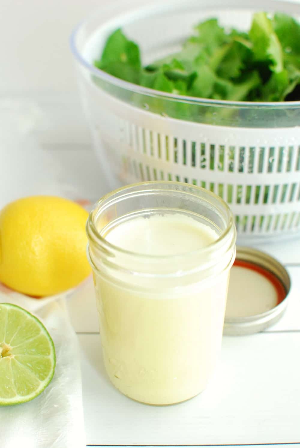 A jar of citrus salad dressing next to a lemon and a bowl of lettuce.