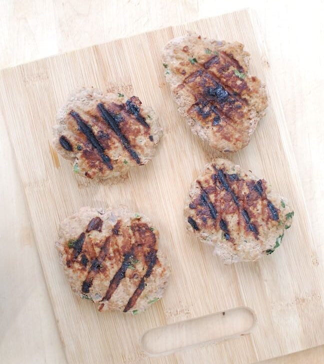 Four cooked burger patties on a wooden cutting board.