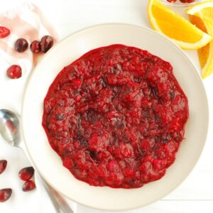 A bowl of lower sugar cranberry sauce next to some fresh orange slices and fresh cranberries.