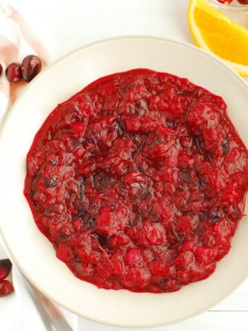 A bowl of lower sugar cranberry sauce next to some fresh orange slices and fresh cranberries.