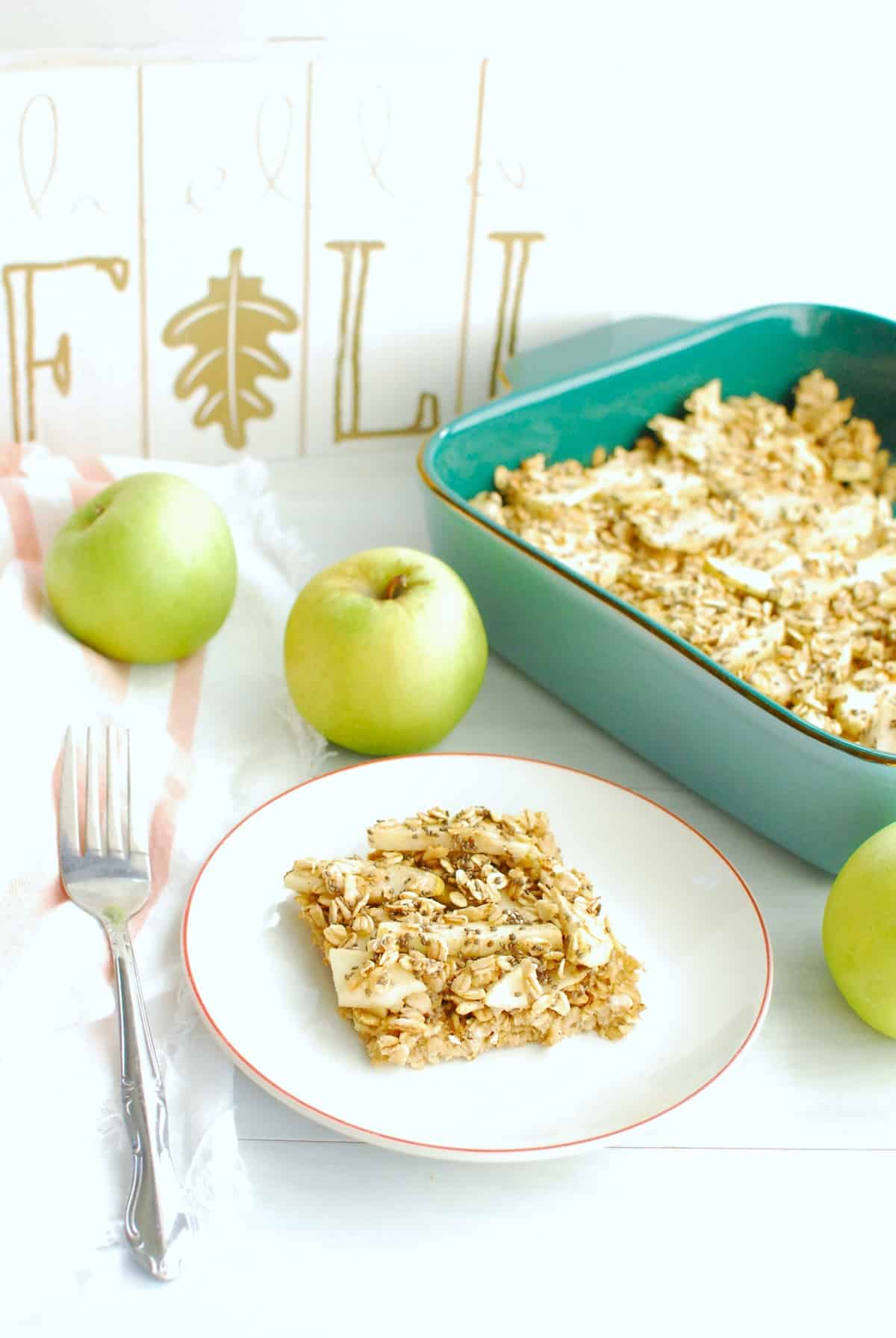 A healthy apple breakfast bar on a white plate next to a fork, napkin, apples, and baking dish.
