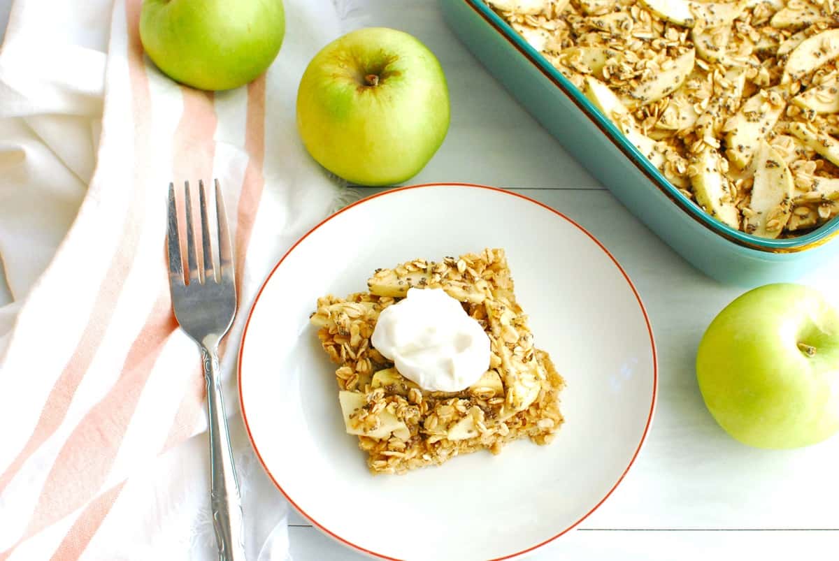 Overhead shot of an apple oatmeal bar topped with Greek yogurt, on a plate next to a napkin and apples.