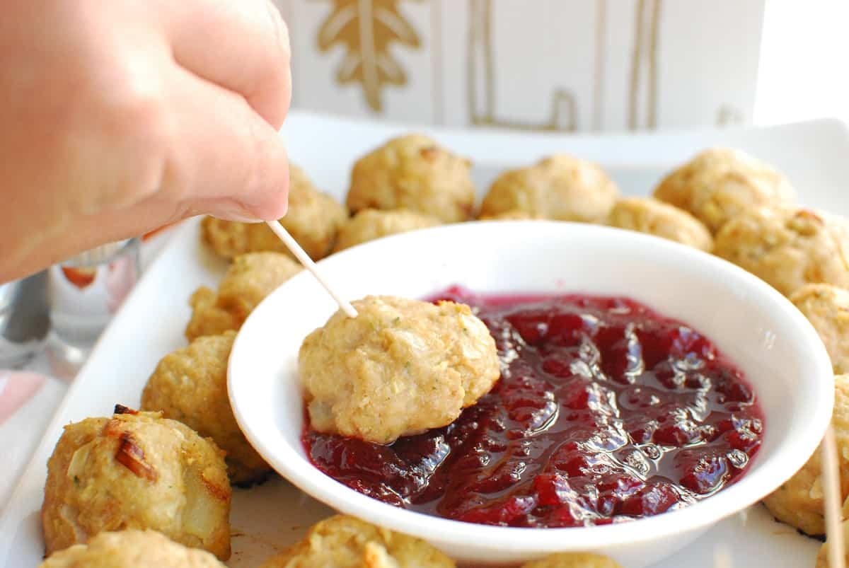 A woman's hand holding a toothpick with a chicken apple meatball, being dipped into cranberry sauce.