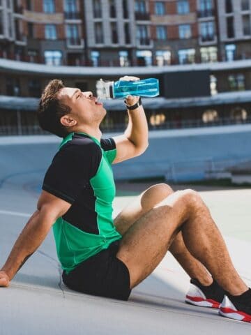 A male runner sitting at an outdoor track drinking water.