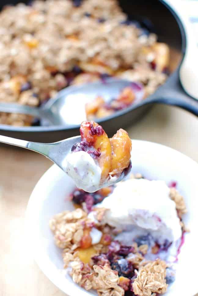 A spoonful of blueberry peach crisp and ice cream held above a bowl.