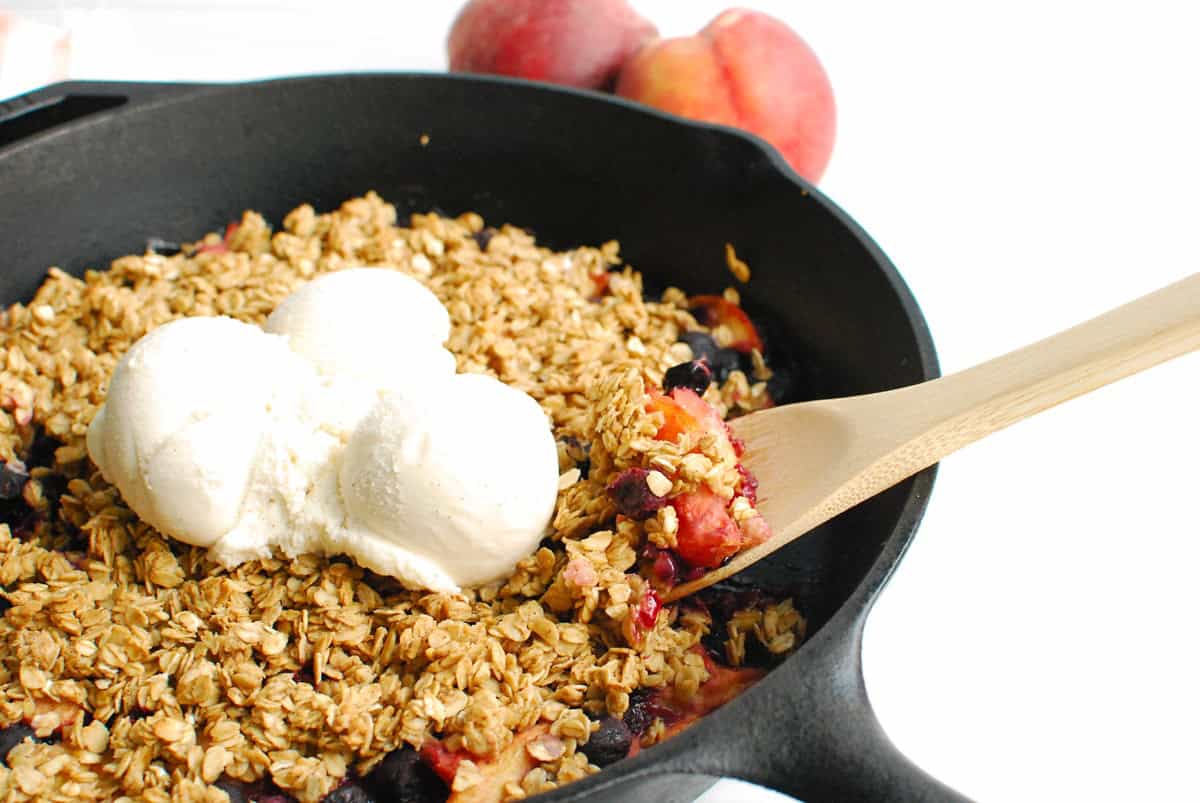 A wooden spoon scooping out some peach blueberry crisp from a cast iron skillet.