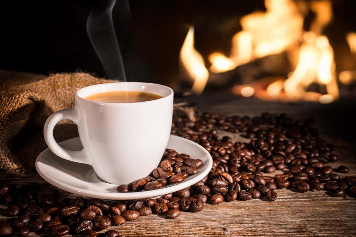 A cup of coffee next to some coffee beans with a fireplace in the background.