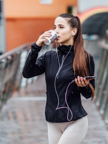 A woman drinking coffee before running, standing outside with headphones on and wearing athletic clothes.