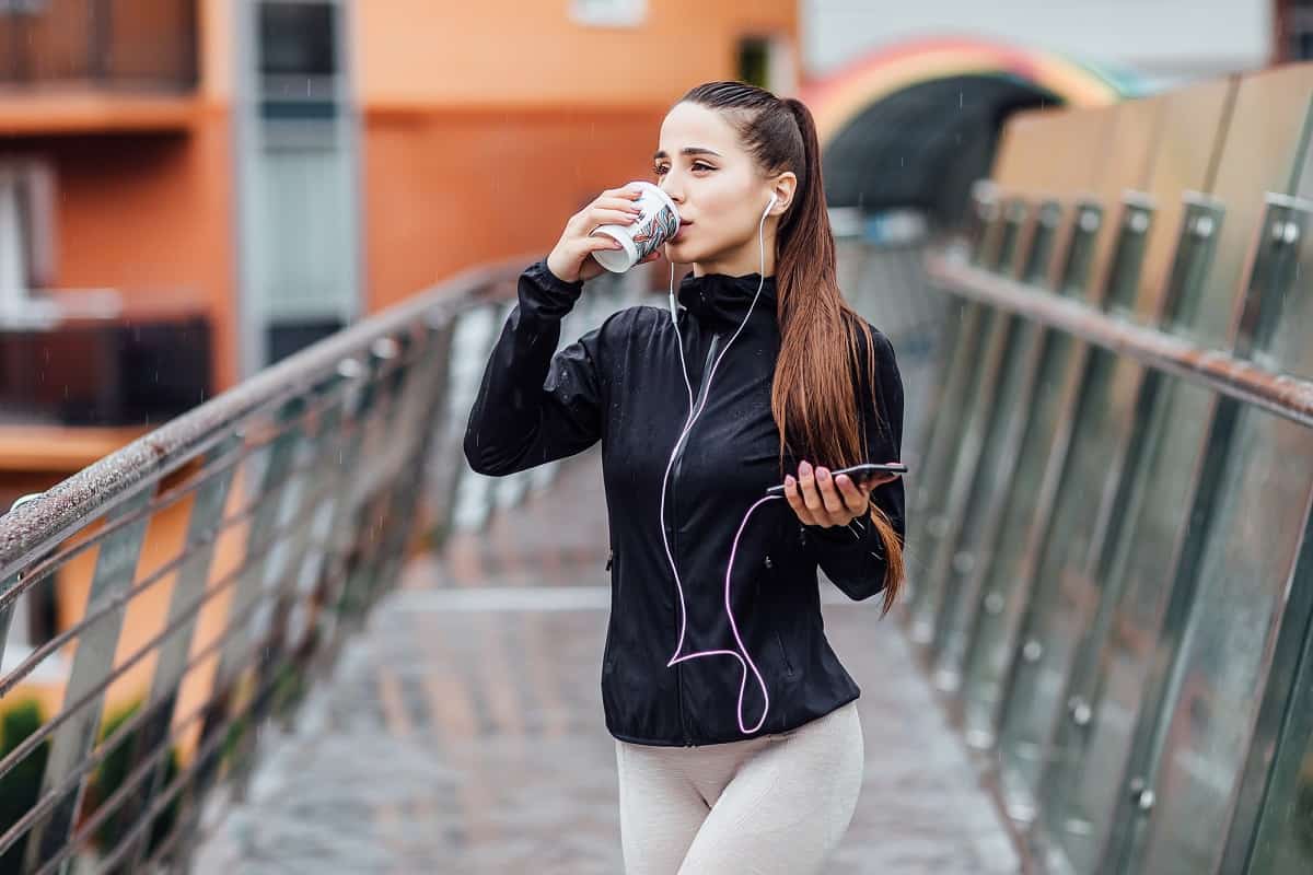 A woman drinking coffee before running, standing outside with headphones on and wearing athletic clothes.
