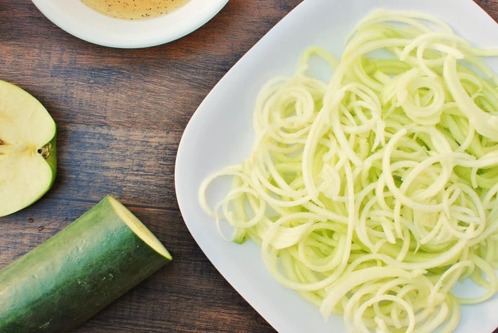 a white plate with cucumber apple salad, next to a piece of a cucumber and a chopped apple