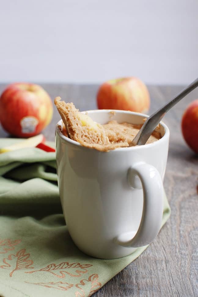 an apple mug cake with a spoon taking a scoop out of it, next to some fresh apples