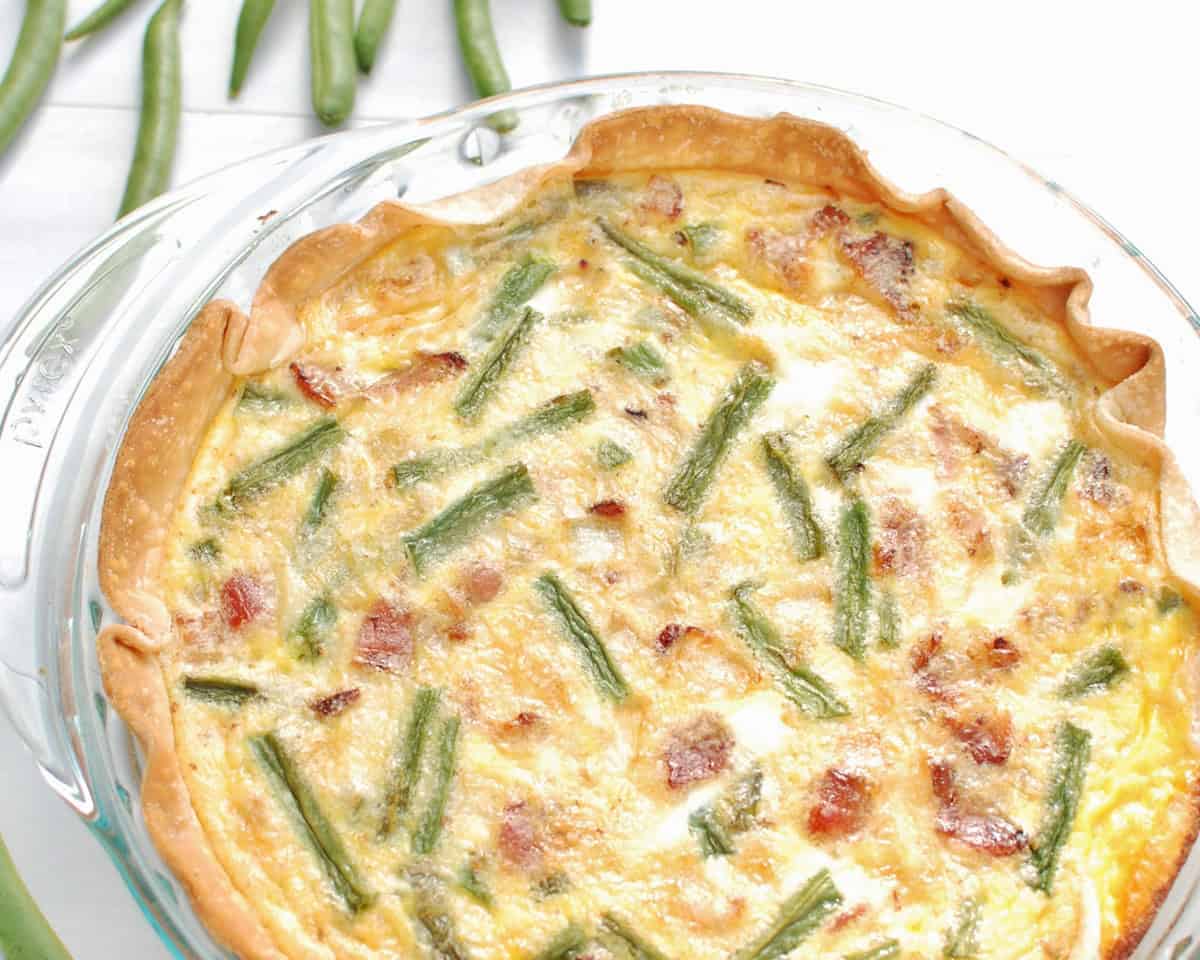 A green bean and bacon quiche in a glass pie dish, next to some fresh green beans.