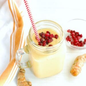 A turmeric banana smoothie next to fresh turmeric, pomegranate arils, and a napkin.