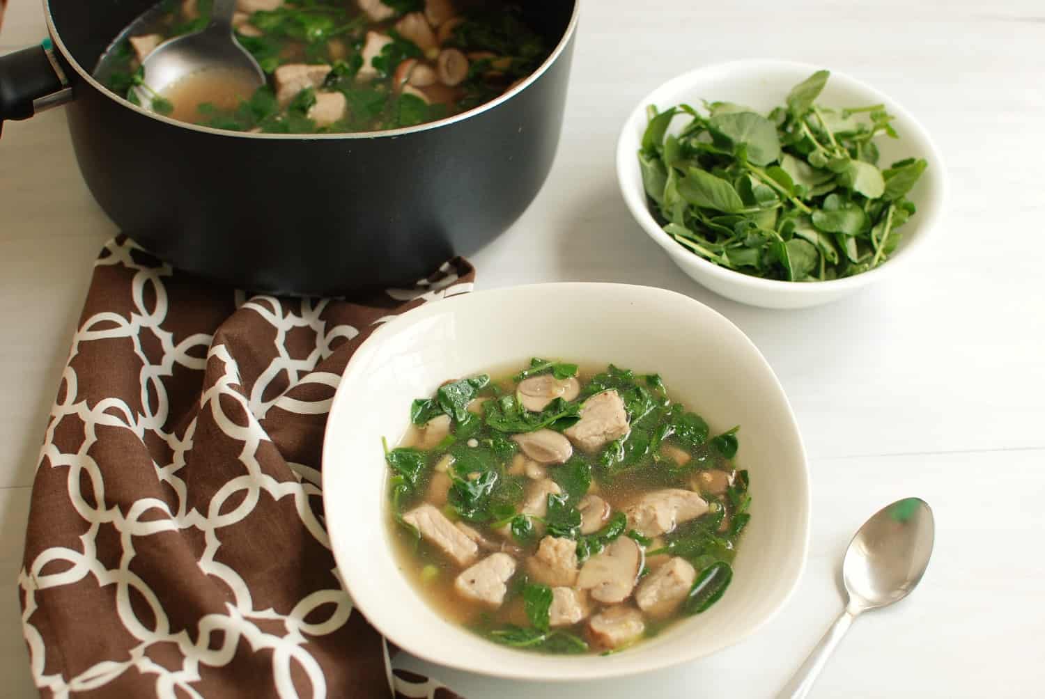 A bowl and pot of pork watercress soup, next to a bowl of watercress