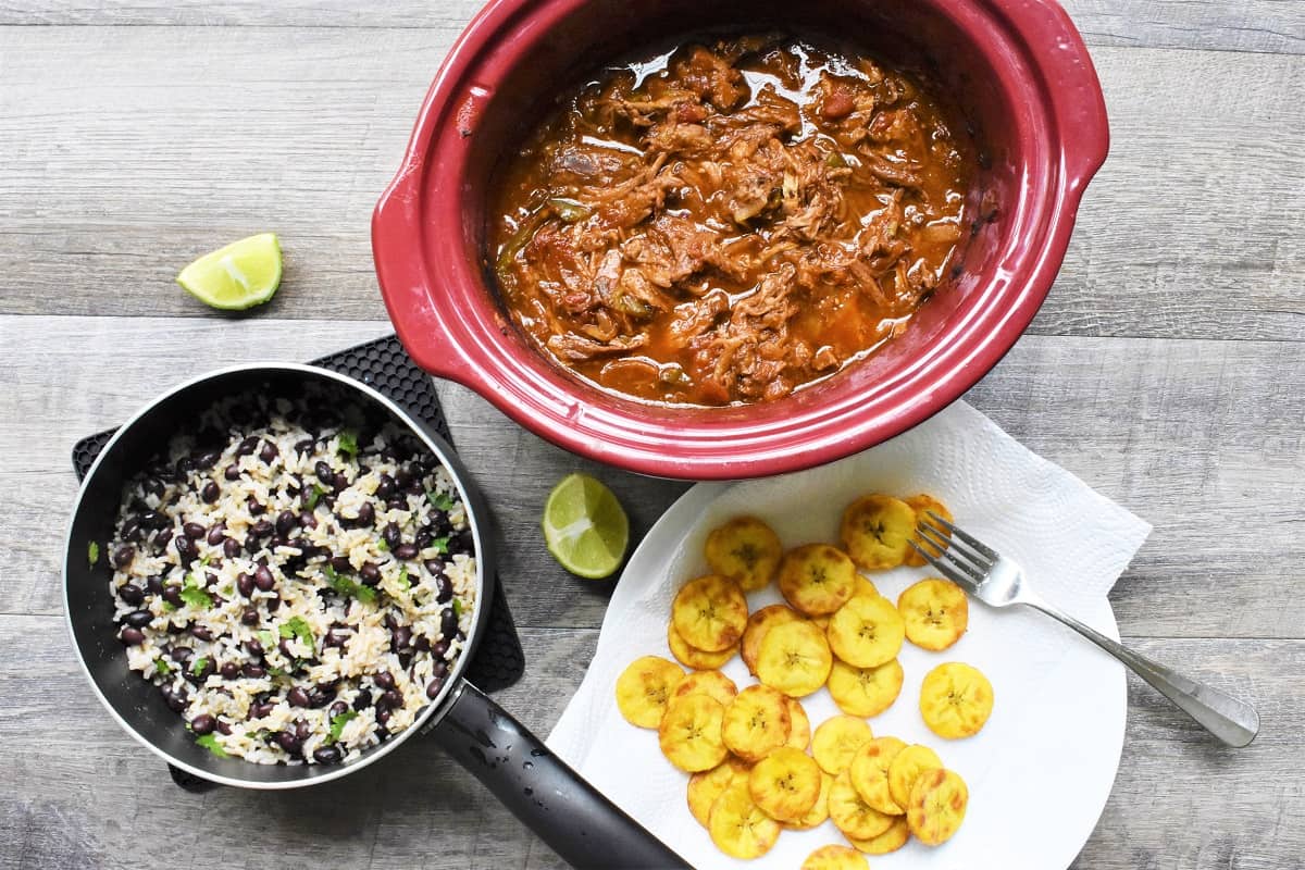 A crockpot with shredded beef, a pot with rice and beans, and a plate with fried plantains.