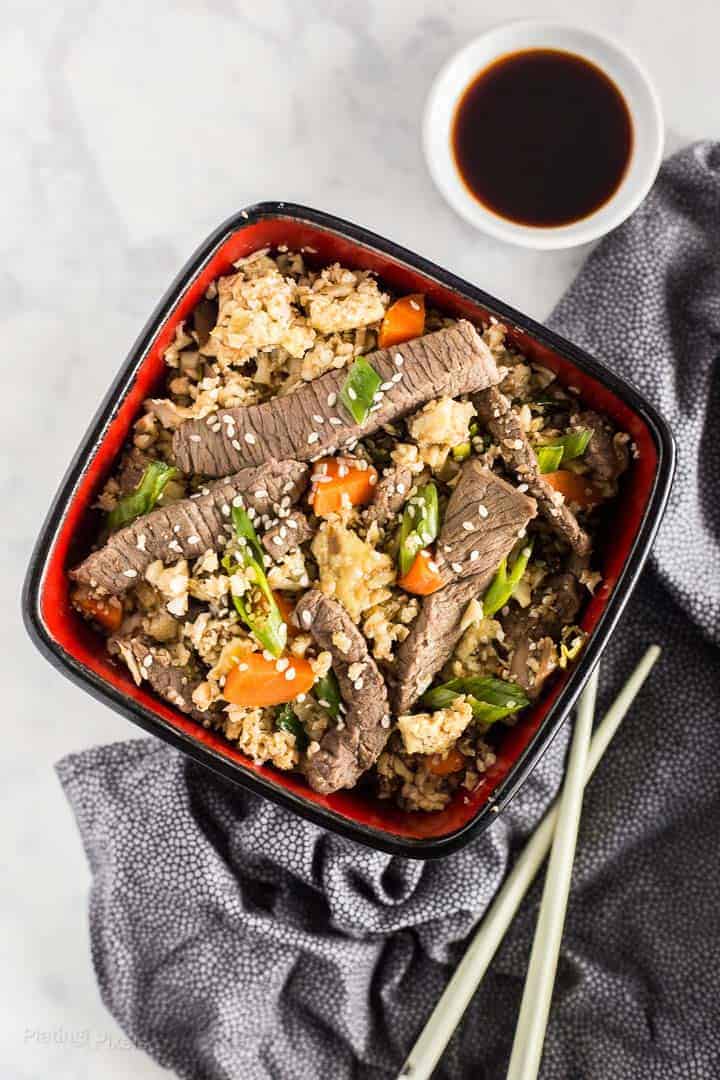 Beef fried rice in a red and black bowl next to a white cup of soy sauce.