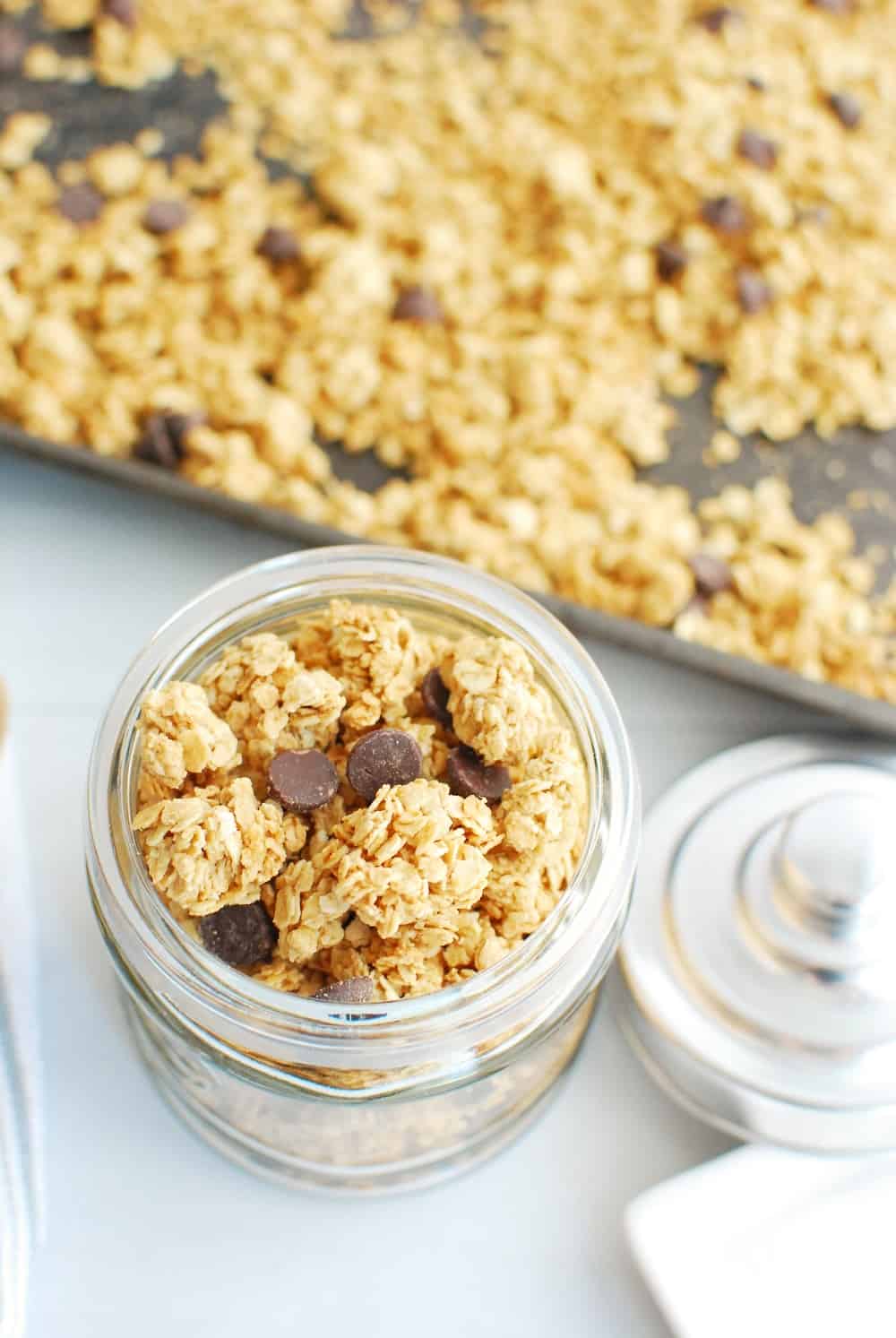 A small jar full of homemade vegan granola next to a large sheet pan of granola