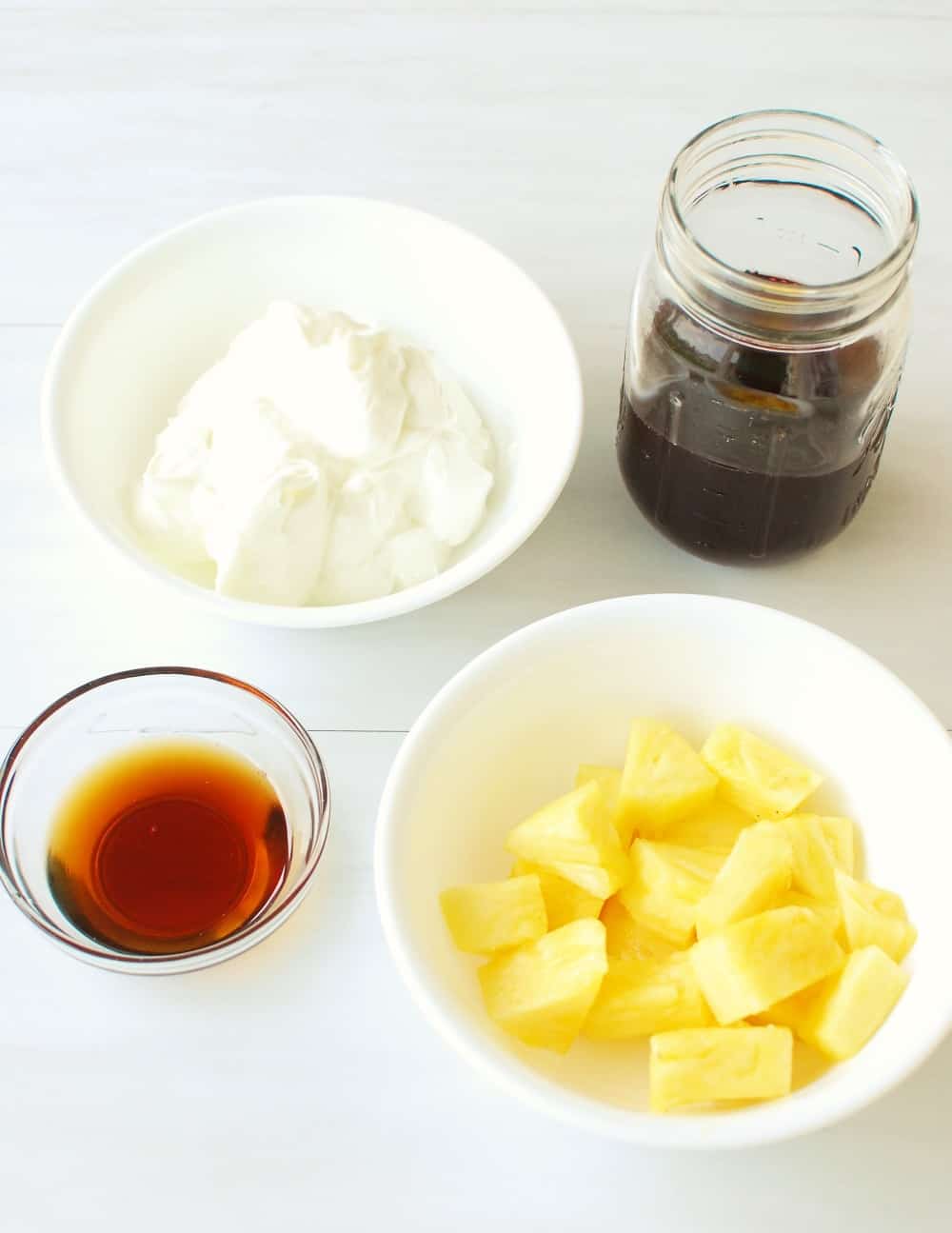 Chopped pineapple in a bowl, maple syrup, a bowl of greek yogurt, and a glass of cherry juice