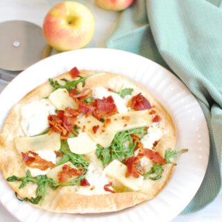 A tortilla pizza topped with arugula, mozzarella, bacon, and apple - on a white plate