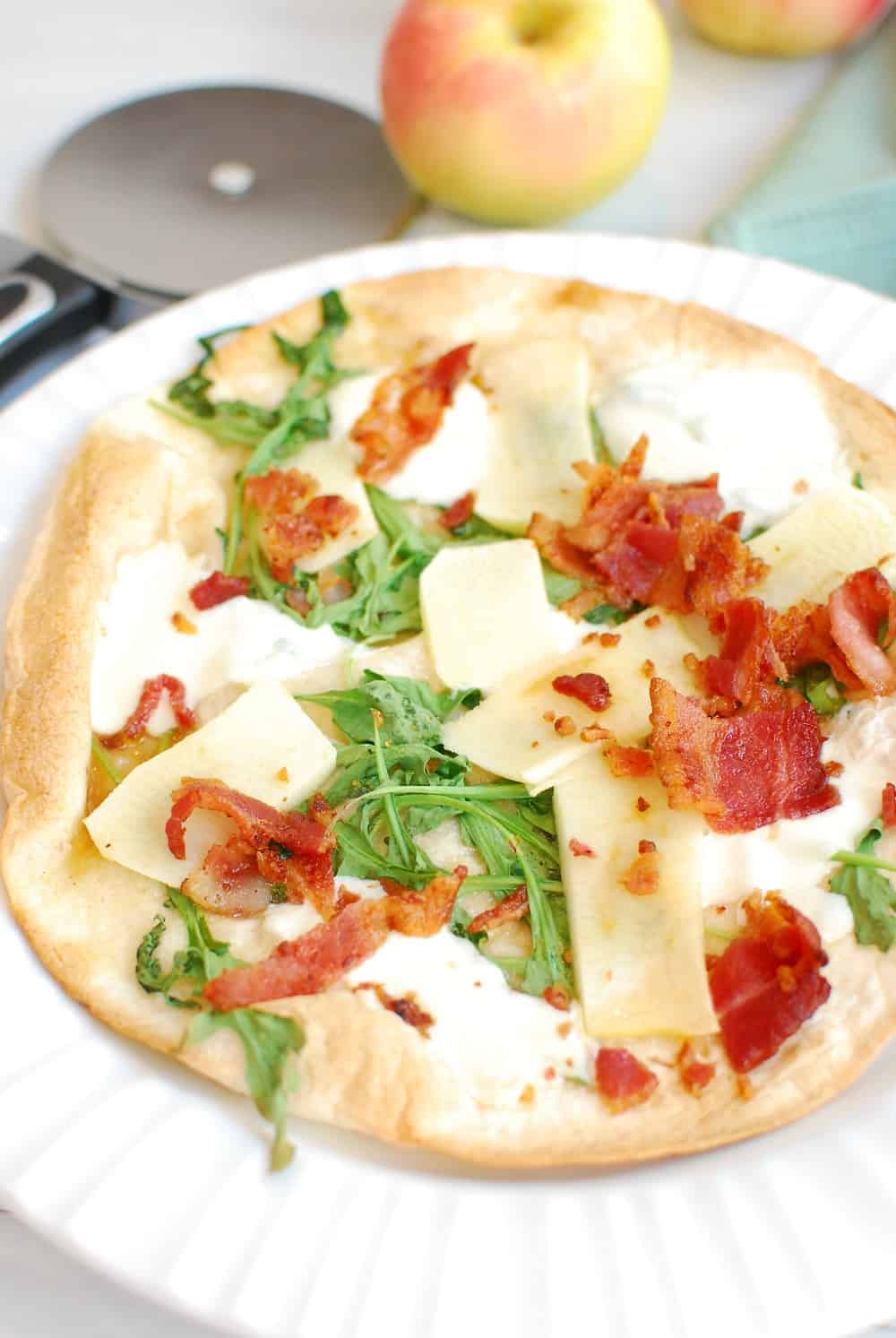 A tortilla pizza on a white plate next to a pizza cutter and an apple