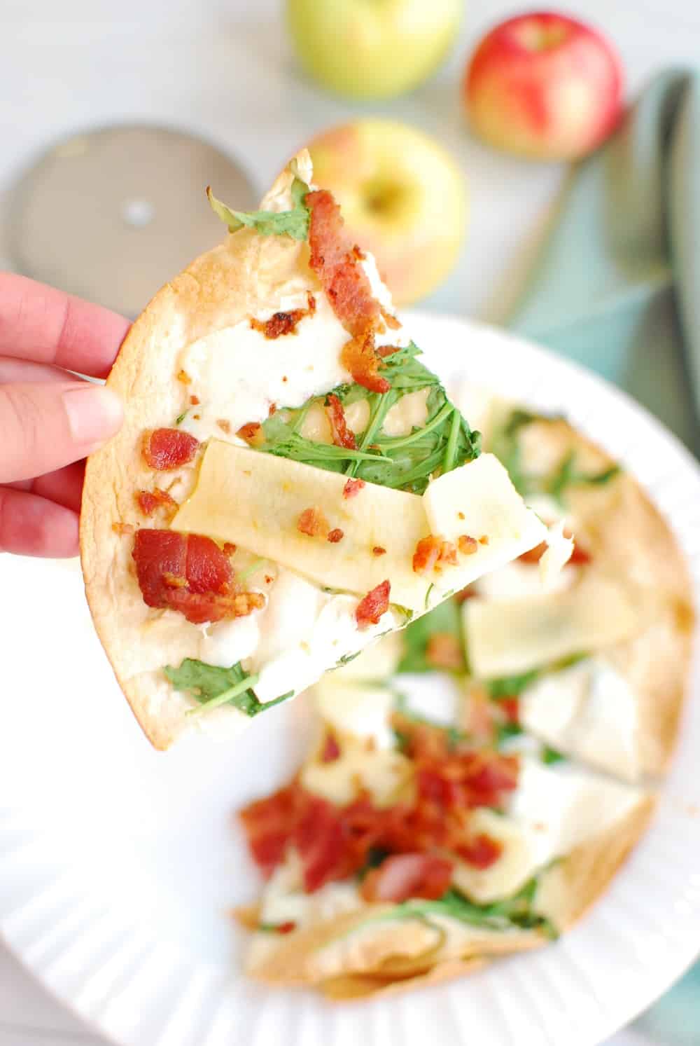 A woman's hand holding a slice of apple tortilla pizza