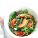 A white bowl full of arugula spinach salad with peaches, pecans, and blue cheese