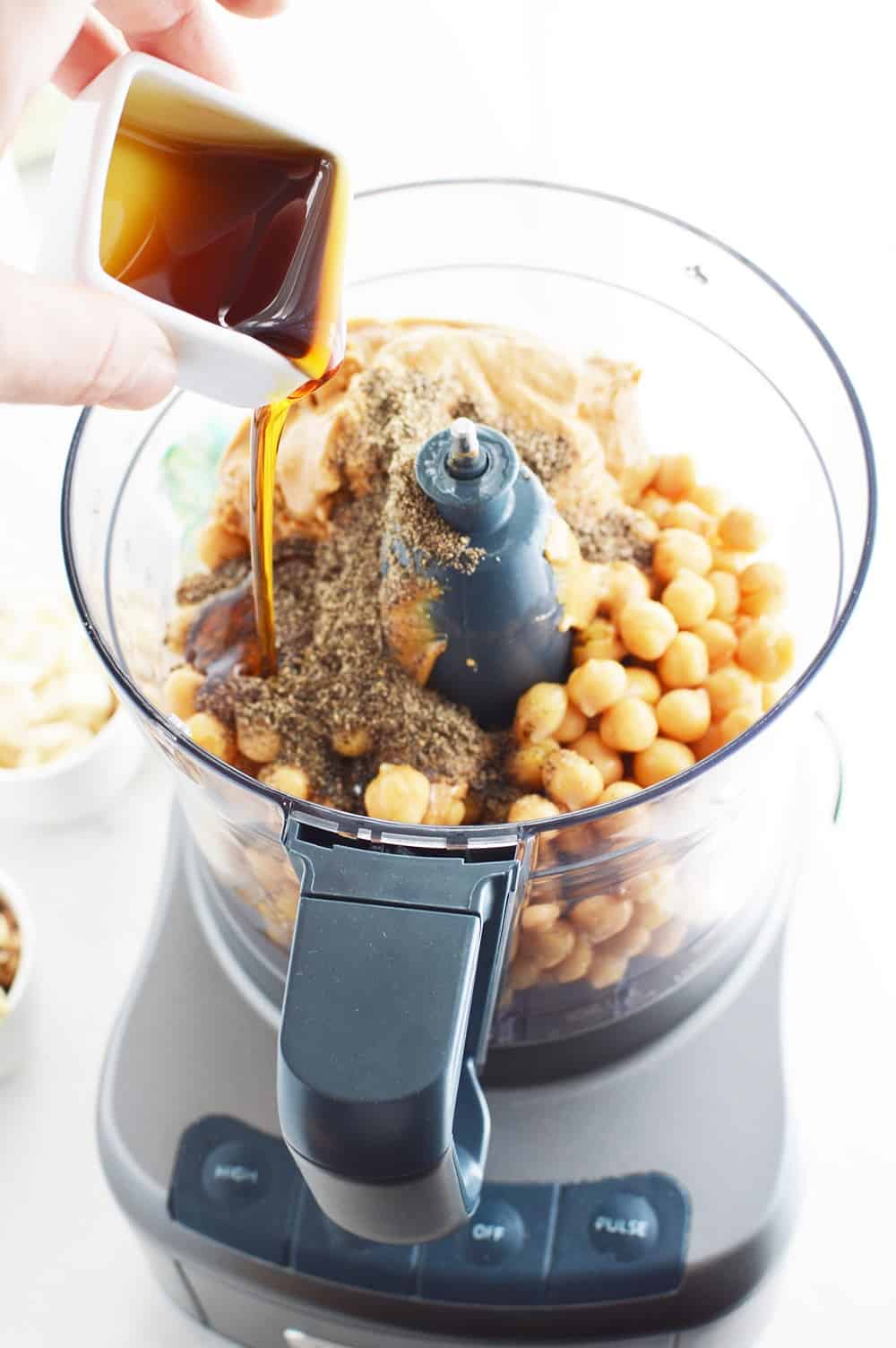 A woman pouring maple syrup into a food processor to make dessert hummus