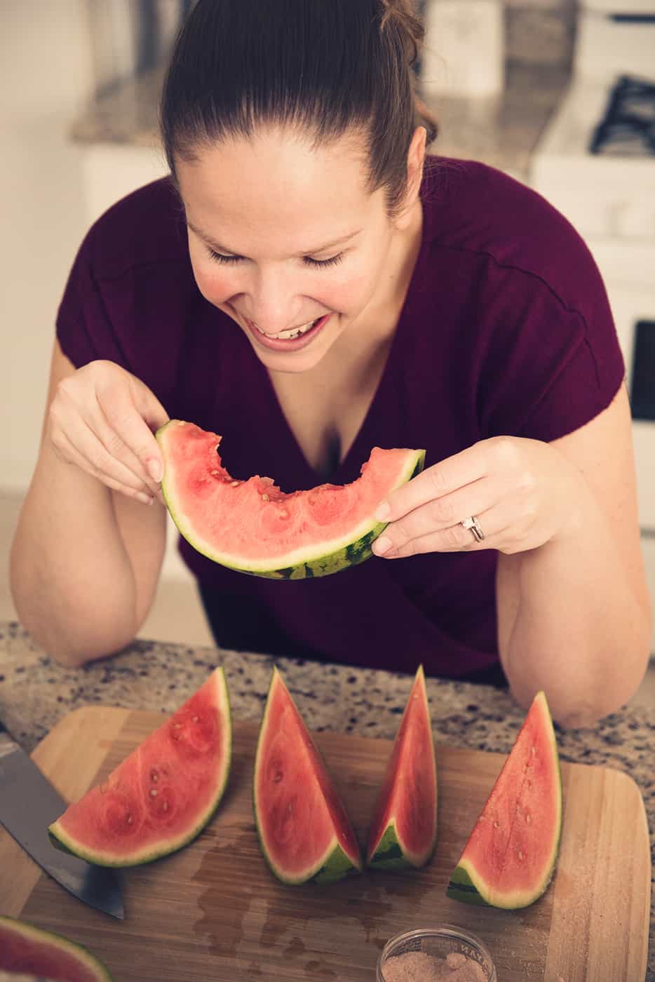 A woman eating a slice of watermelon at the kitchen counter