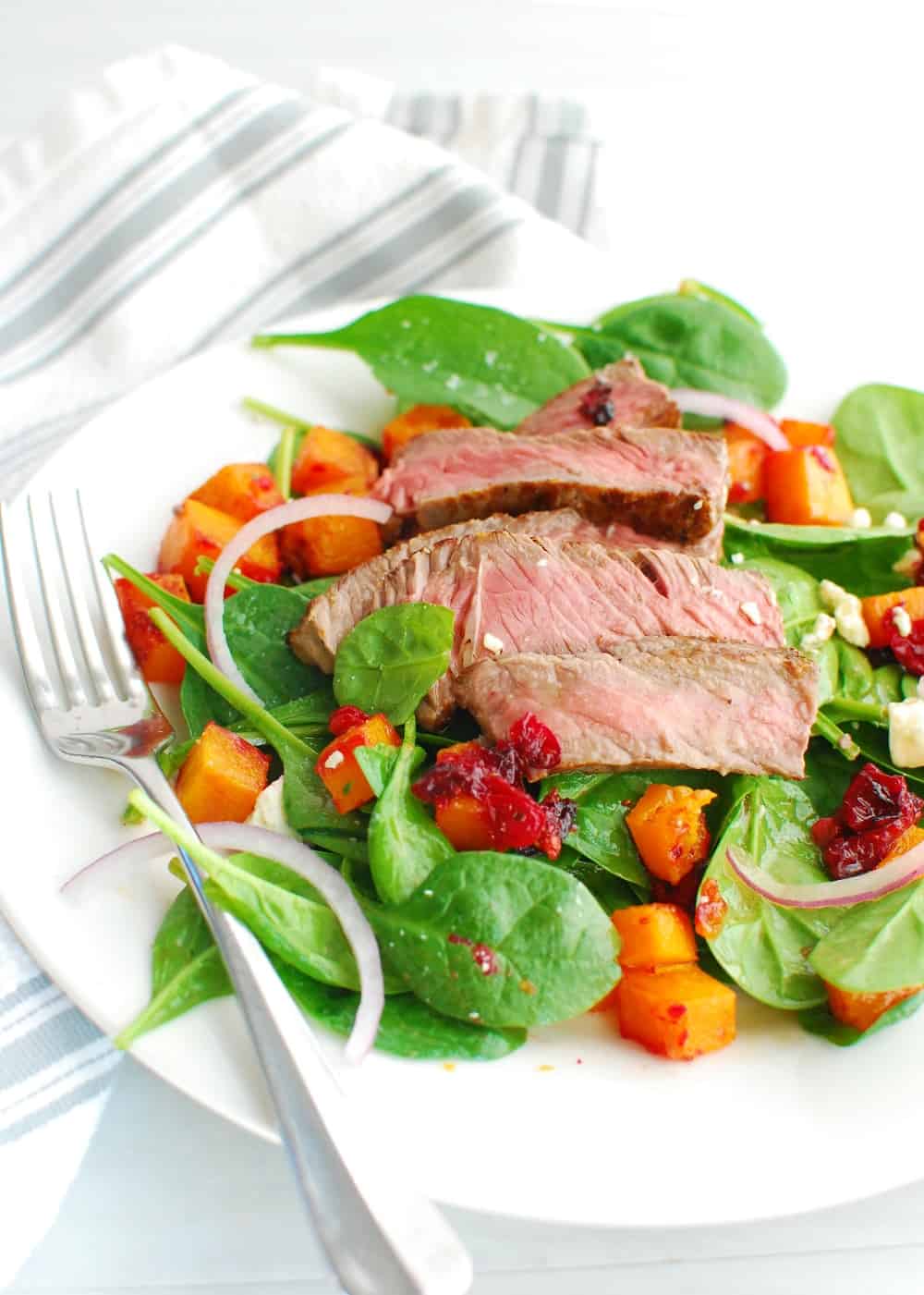 A plate full of steak and spinach salad topped with squash, cranberries and blue cheese