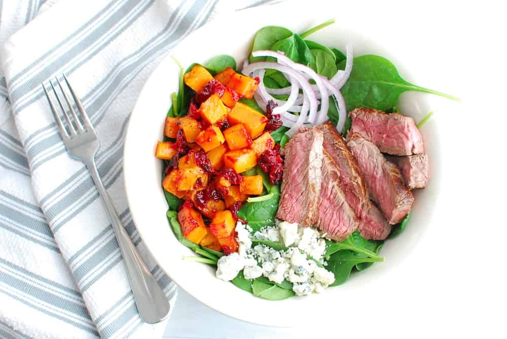 A steak and blue cheese salad with butternut squash and cranberries in a white bowl