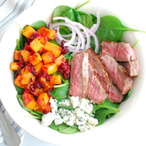 A steak salad with butternut squash, cranberries, blue cheese, and red onion.
