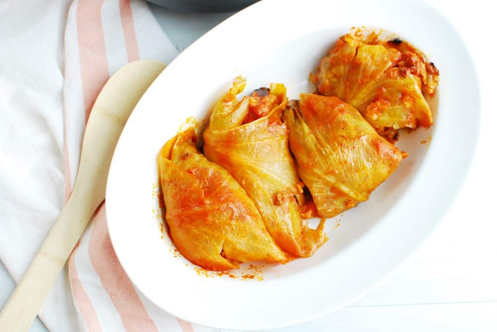 Several stuffed cabbage rolls in a white dish next to a napkin and wooden spoon.