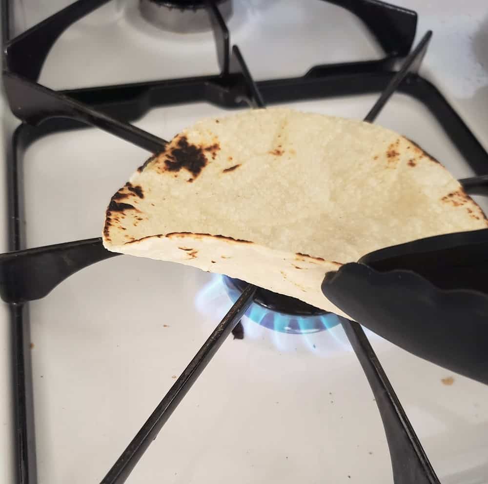 a corn tortilla on top of the stovetop, about to be flipped with tongs