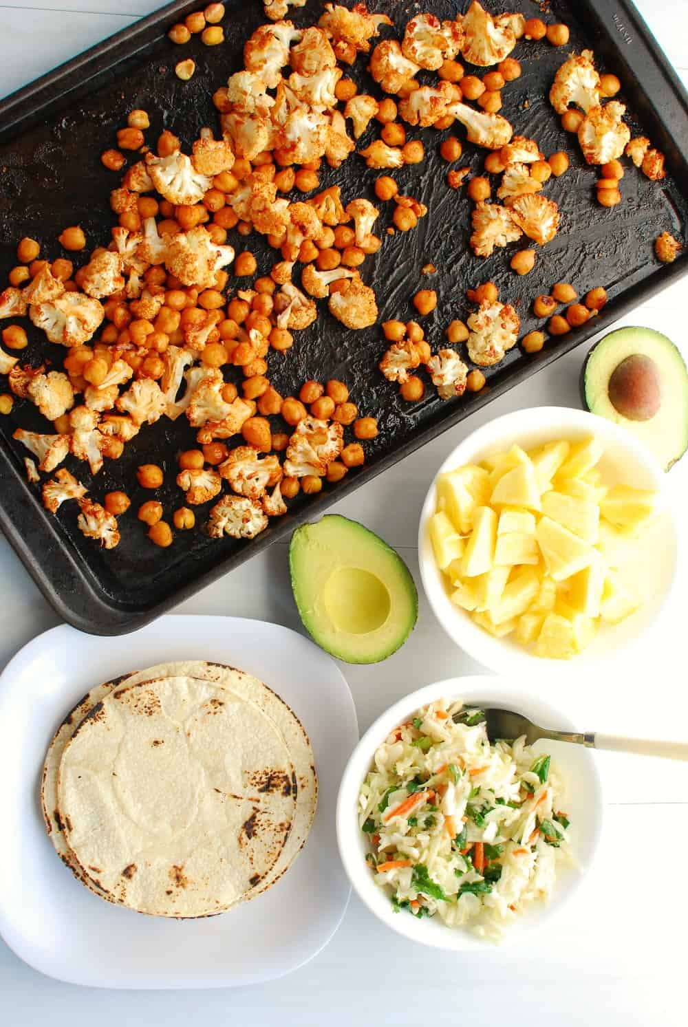 a pan of bbq cauliflower and chickpeas next to a plate with corn tortillas, a bowl with pineapple, an avocado, and a bowl of cabbage slaw