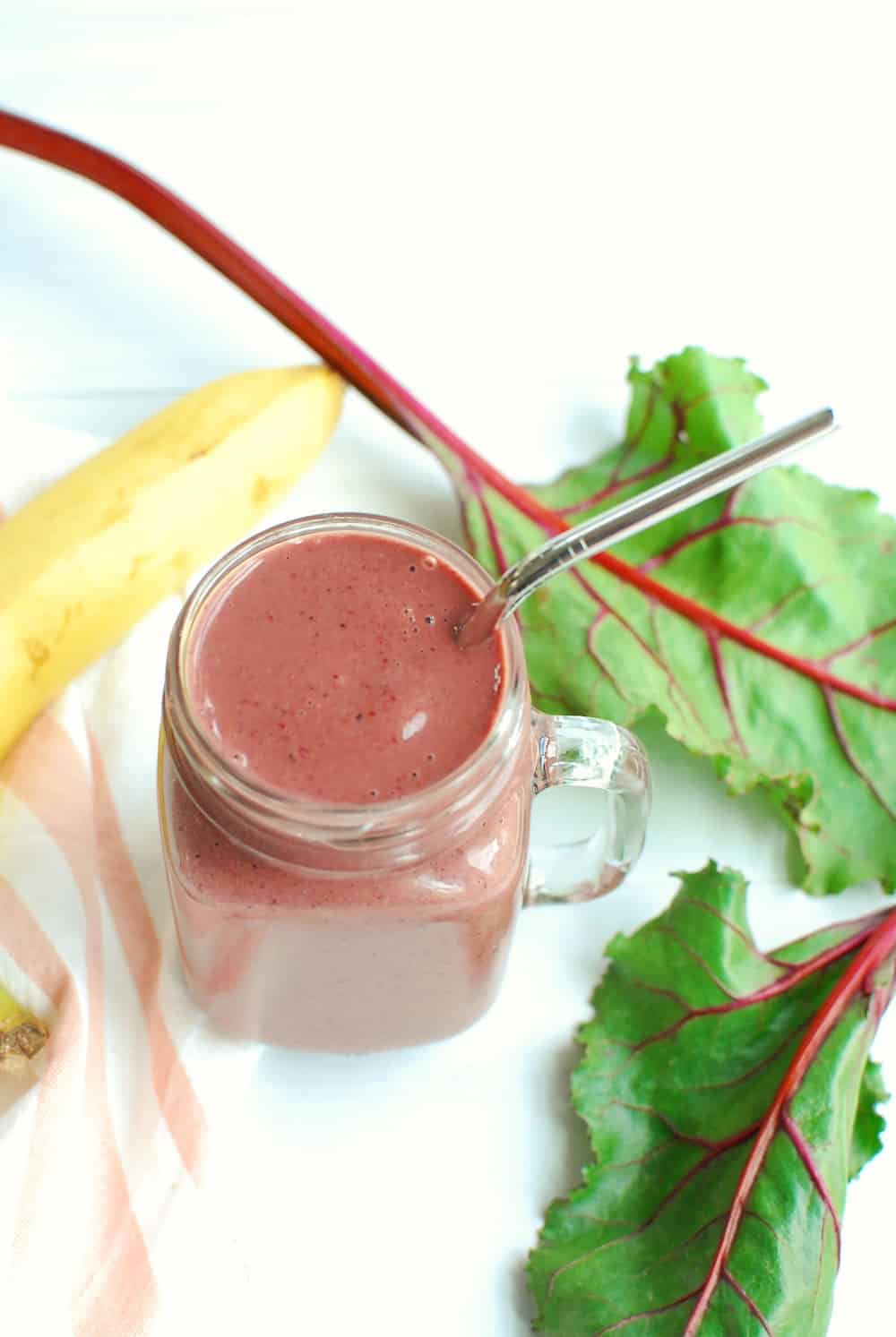 a mason jar with a banana, berry, and beet green smoothie in it, next to a banana and some raw fresh beet greens